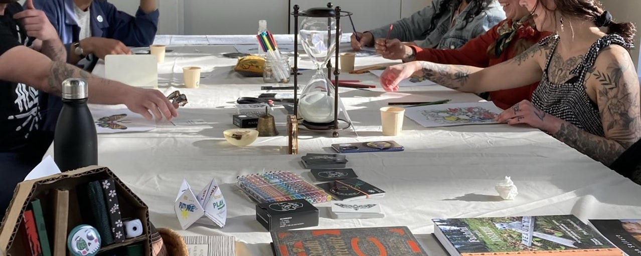 A group of people gathered at a table colouring and playing Death Conversation Game. An Hourglass sits in the center of the table.